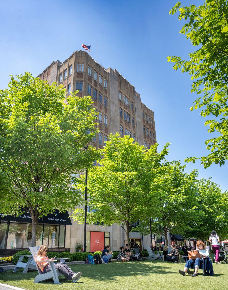 A central green with people lounging on the grass and in Adirondack chairs with boutique shops in the background